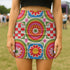 A vibrant bodycon mini skirt featuring a structured grid of retro geometric medallions. The pattern includes concentric circles in red, yellow, and blue, stylized daisy flower motifs, and red and white checkered squares. The skirt has a high-waist elastic band and a smooth, matte performance knit texture. The background is a neutral off-white. This maximalist design utilizes saturated primary colors to create a 70s-inspired psychedelic aesthetic. No purple or navy is present in the print.