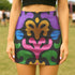 A high-waisted bodycon mini skirt for women shown in two fabric options. The skirt features a vibrant, colorful, symmetrical abstract print with psychedelic, groovy, baroque-inspired shapes in purple, pink, gold, green, and blue on a black background. One version is a smooth, stretchy signature performance knit that is UV reactive and glows under black light. The second version is a glitter knit with a metallic, shimmery foil finish. The mid-thigh length skirt has a tight, curve-hugging fit.