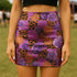 A vibrant, curve-hugging bodycon mini skirt featuring a maximalist mix of orange and purple checkerboard, black leopard print, and realistic rose florals in purple and orange tones. The high-waisted, mid-thigh cut is designed for a confident fit. Made from a durable performance knit fabric with two finish options: a signature UV reactive version that glows under blacklight for festivals and a dazzling glitter knit with reflective silver foil for a party-ready shimmer.