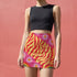 Form-fitting mini skirt featuring a vibrant, retro-inspired maximalist pattern. The design showcases a diagonal hot pink band filled with white and yellow daisies, contrasting against a bold background of wavy, abstract red and yellow flame-like stripes. The bottom edge includes curved color blocks of green and orange. Made of a smooth, stretchy fabric with a mid-rise elastic waistband, hitting at mid-thigh. Available in both a matte black-light reactive finish and a metallic glitter finish.