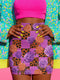 A vibrant, curve-hugging bodycon mini skirt featuring a maximalist mix of orange and purple checkerboard, black leopard print, and realistic rose florals in purple and orange tones. The high-waisted, mid-thigh cut is designed for a confident fit. Made from a durable performance knit fabric with two finish options: a signature UV reactive version that glows under blacklight for festivals and a dazzling glitter knit with reflective silver foil for a party-ready shimmer.