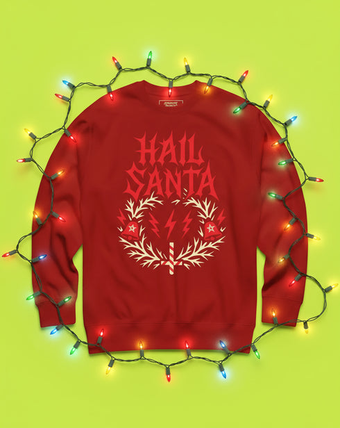 A unisex crewneck sweatshirt shown in red, green, and blue color options. The graphic features "HAIL SANTA" in a red, jagged, heavy metal-style font. The text is framed by a cream-colored wreath made of thorny branches. Inside the wreath are two red lightning bolts and two red bells with cream-colored pentagrams. An inverted candy cane cross sits at the bottom. This alternative holiday sweater has fleece texture, ribbed cuffs, and a classic fit, designed for goth, punk, and metalhead aesthetics.