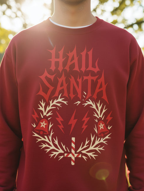 A unisex crewneck sweatshirt shown in red, green, and blue color options. The graphic features "HAIL SANTA" in a red, jagged, heavy metal-style font. The text is framed by a cream-colored wreath made of thorny branches. Inside the wreath are two red lightning bolts and two red bells with cream-colored pentagrams. An inverted candy cane cross sits at the bottom. This alternative holiday sweater has fleece texture, ribbed cuffs, and a classic fit, designed for goth, punk, and metalhead aesthetics.