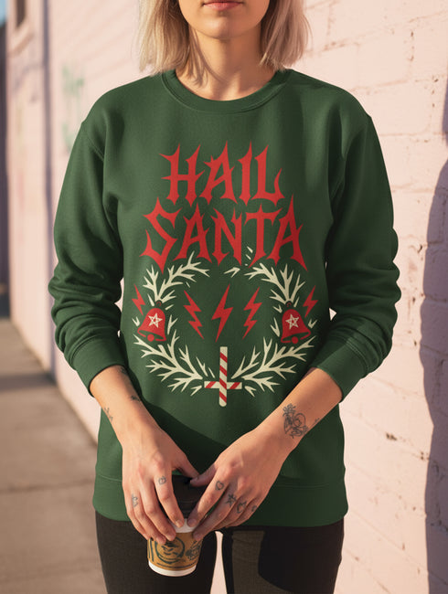 A unisex crewneck sweatshirt shown in red, green, and blue color options. The graphic features "HAIL SANTA" in a red, jagged, heavy metal-style font. The text is framed by a cream-colored wreath made of thorny branches. Inside the wreath are two red lightning bolts and two red bells with cream-colored pentagrams. An inverted candy cane cross sits at the bottom. This alternative holiday sweater has fleece texture, ribbed cuffs, and a classic fit, designed for goth, punk, and metalhead aesthetics.