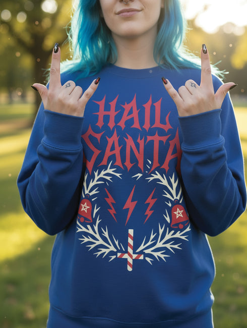 A unisex crewneck sweatshirt shown in red, green, and blue color options. The graphic features "HAIL SANTA" in a red, jagged, heavy metal-style font. The text is framed by a cream-colored wreath made of thorny branches. Inside the wreath are two red lightning bolts and two red bells with cream-colored pentagrams. An inverted candy cane cross sits at the bottom. This alternative holiday sweater has fleece texture, ribbed cuffs, and a classic fit, designed for goth, punk, and metalhead aesthetics.