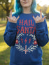 A unisex crewneck sweatshirt shown in red, green, and blue color options. The graphic features "HAIL SANTA" in a red, jagged, heavy metal-style font. The text is framed by a cream-colored wreath made of thorny branches. Inside the wreath are two red lightning bolts and two red bells with cream-colored pentagrams. An inverted candy cane cross sits at the bottom. This alternative holiday sweater has fleece texture, ribbed cuffs, and a classic fit, designed for goth, punk, and metalhead aesthetics.
