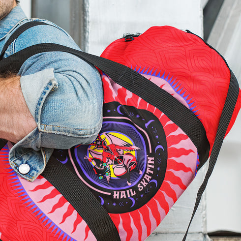 Red cylindrical roller skating duffle bag with black straps, featuring a prominent circular emblem with a colorful Baphomet design and "Hail Skatin" text. Includes mesh side pocket and adjustable padded shoulder strap.