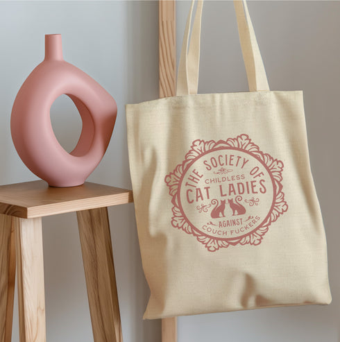 Election 2024 Canvas tote bag featuring a circular design with two cats and text "The Society of Childless Cat Ladies Against Couch Fuckers" in burgundy on a natural or black.