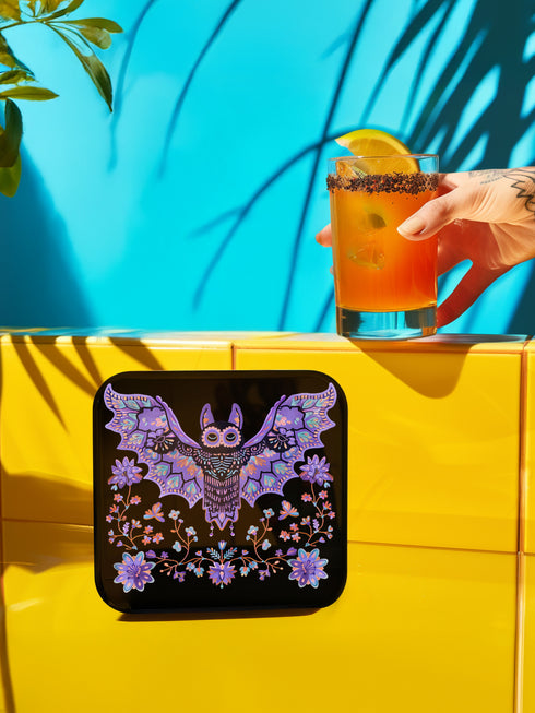 Square coasters featuring lavender-purple bats with folk art floral designs against black backgrounds. Pastel goth drink coasters, kawaii bat home decor, whimsigoth barware accessories, gothic floral table protection for weird home aesthetics.