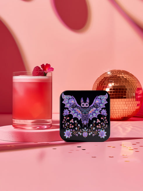 Square coasters featuring lavender-purple bats with folk art floral designs against black backgrounds. Pastel goth drink coasters, kawaii bat home decor, whimsigoth barware accessories, gothic floral table protection for weird home aesthetics.