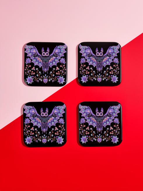 Square coasters featuring lavender-purple bats with folk art floral designs against black backgrounds. Pastel goth drink coasters, kawaii bat home decor, whimsigoth barware accessories, gothic floral table protection for weird home aesthetics.