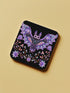 Square coasters featuring lavender-purple bats with folk art floral designs against black backgrounds. Pastel goth drink coasters, kawaii bat home decor, whimsigoth barware accessories, gothic floral table protection for weird home aesthetics.