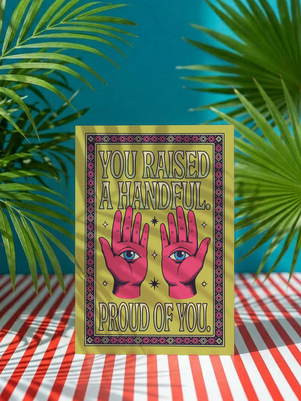 Funny Mother’s Day card in a 5x7 size featuring a vibrant chartreuse green background. The centralized design shows two pink hands with an electric blue eye in each palm, surrounded by cream stars. The retro bubble text reads "YOU RAISED A HANDFUL. PROUD OF YOU." A plum and pink geometric diamond border frames the card. This high-fidelity print mimics a 70s psychedelic art style with a flat matte or silk finish. A white sealable envelope is included with the handmade card.