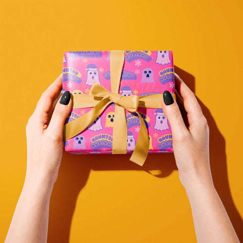Bright pink christmas wrapping paper covered in cute ghost characters wearing Santa hats, with "Haunted Holiday" and "Boo Humbug" text, and snowflake accents.
