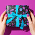 Spooky birthday wrapping paper with neon pink skulls, blue coffins, eyeballs, and bats on black background. Vibrant and gothic design for unique gift presentation.