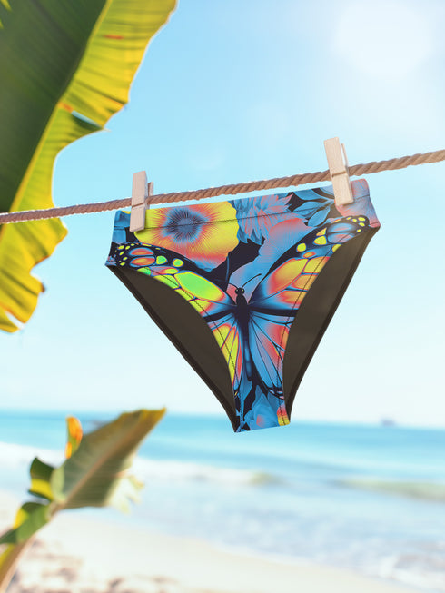 High-waisted bikini bottom with a bold psychedelic butterfly design in neon shades of orange, yellow, blue, and pink on a black background. Features cheeky fit and double-layered fabric, lying flat against a turquoise swimming pool backdrop. Perfect for rave swimwear, Y2K beach looks, and festival-ready poolside outfits. Vibrant and colorful statement swimwear for bold summer style.