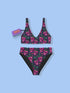 Two-piece bikini set featuring a bold pastel goth design with neon pink skulls wearing green bows, surrounded by multicolored stars on a deep black background. The set includes a plunging V-neck bikini top with wide straps and high-waisted bottoms for a flattering retro fit. Made of stretchy, soft material with a smooth black lining. This skull print swimsuit is ideal for alternative, edgy summer style with a playful punk twist.