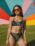 High-waisted bikini bottom featuring a holographic snake scale print with neon rainbow tones on a sleek black background. Iridescent hexagon patterns add a futuristic edge. Cheeky fit with a double-layered finish, styled poolside against a bright turquoise water backdrop. Perfect for rave-ready swim looks, alternative beachwear, and bold festival swimwear with a Y2K cyberpunk twist.
