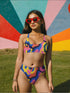 High-waisted bikini bottom with a vibrant wavy print in rainbow hues of pink, blue, yellow, orange, and navy on a black base. Features cheeky fit and double-layered fabric, styled over a bright turquoise pool for a sun-drenched summer look. Perfect for maximalist beachwear, retro pool parties, and colorful rave-inspired swim fashion.