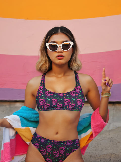 Two-piece bikini set featuring a bold pastel goth design with neon pink skulls wearing green bows, surrounded by multicolored stars on a deep black background. The set includes a plunging V-neck bikini top with wide straps and high-waisted bottoms for a flattering retro fit. Made of stretchy, soft material with a smooth black lining. This skull print swimsuit is ideal for alternative, edgy summer style with a playful punk twist.