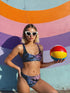Model wearing a high-waisted pastel goth bikini set featuring a black bat and spiderweb print over an ombré pink and purple background. The two-piece swimsuit includes a scoop neck top and high-rise bottoms, styled outdoors in bright festival and poolside settings. Made of recycled fabric, soft, stretchy, and UV resistant. Available in XS to 3X. Aesthetic is gothic, spooky cute, and Y2K inspired.