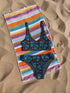 High-waisted bikini set featuring an all-over pattern of neon blue skulls with pink bows, pastel stars, and purple cherries on a dark olive green background. The top has a scoop neckline with wide straps, and the bottoms offer full coverage. A bold, punk-inspired swim look perfect for alternative fashion lovers and dopamine dressers.