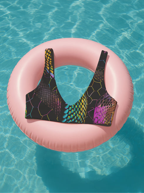 Sports-bra-style bikini top with wide straps and scoop neckline, featuring a vibrant holographic snake print in neon yellow, pink, purple, and blue tones over black. Hexagon pattern overlays add a cyber-sleek finish. Floating above a turquoise pool, this statement swim top brings futuristic Y2K energy to any swimwear collection. Ideal for alt baddies, rave queens, and dopamine dressers.