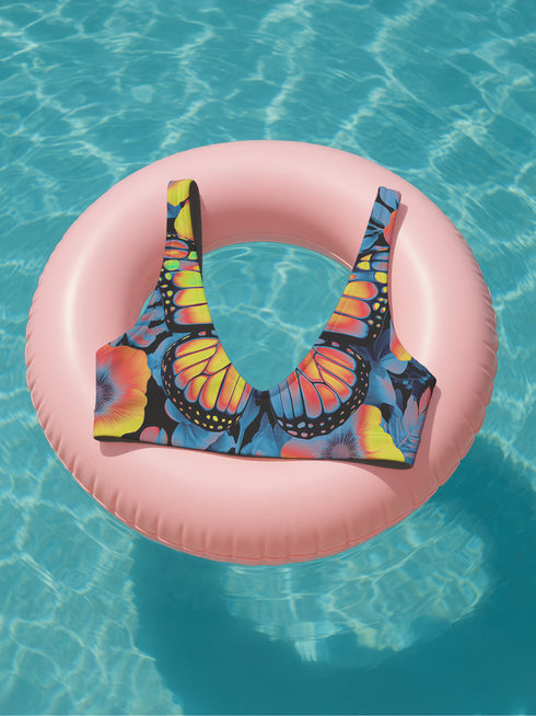 Butterfly-print bikini top with a vivid psychedelic monarch wing design in neon yellow, orange, and pink on a black base. Features a scoop neck, sports bra-style silhouette, and wide shoulder straps. Floating over a bright turquoise swimming pool, this swim top blends Y2K rave energy with bold festival fashion. Perfect for alt-girls, beach rebels, and dopamine dressers craving color-drenched swimwear.