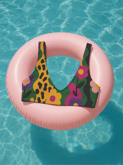 Scoop-neck bikini top with thick straps and a vibrant retro flower print in yellow, pink, green, and black. Features a secure, supportive sports-bra style fit. Shown above a bright turquoise pool, this top channels a maximalist 70s energy with bold color blocking and playful floral design. Ideal for statement swimwear, retro summer fashion, and festival season looks.