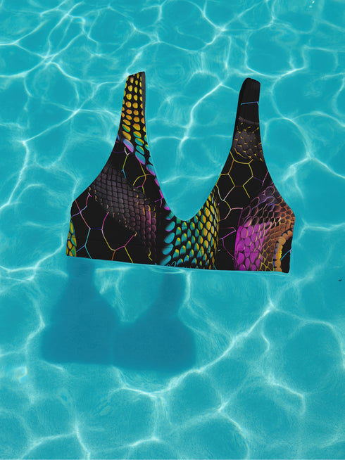 Sports-bra-style bikini top with wide straps and scoop neckline, featuring a vibrant holographic snake print in neon yellow, pink, purple, and blue tones over black. Hexagon pattern overlays add a cyber-sleek finish. Floating above a turquoise pool, this statement swim top brings futuristic Y2K energy to any swimwear collection. Ideal for alt baddies, rave queens, and dopamine dressers.
