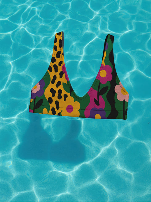 Scoop-neck bikini top with thick straps and a vibrant retro flower print in yellow, pink, green, and black. Features a secure, supportive sports-bra style fit. Shown above a bright turquoise pool, this top channels a maximalist 70s energy with bold color blocking and playful floral design. Ideal for statement swimwear, retro summer fashion, and festival season looks.