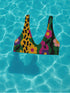 Scoop-neck bikini top with thick straps and a vibrant retro flower print in yellow, pink, green, and black. Features a secure, supportive sports-bra style fit. Shown above a bright turquoise pool, this top channels a maximalist 70s energy with bold color blocking and playful floral design. Ideal for statement swimwear, retro summer fashion, and festival season looks.