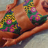 Scoop-neck bikini top with thick straps and a vibrant retro flower print in yellow, pink, green, and black. Features a secure, supportive sports-bra style fit. Shown above a bright turquoise pool, this top channels a maximalist 70s energy with bold color blocking and playful floral design. Ideal for statement swimwear, retro summer fashion, and festival season looks.