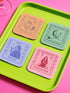"A set of four square coasters featuring humorous tarot card designs in pastel colors. The set includes a pink coaster titled 'High Priestess of Pour Decisions' showing a witch holding a goblet, a purple coaster titled 'Death of My Liver' depicting a skeleton holding a martini, an orange coaster titled 'The Lovers of Bad Idea Texts' featuring two hands toasting, and a green coaster titled 'The Hangover of Next-Day Regret' showing a skeleton falling. Glossy finish.