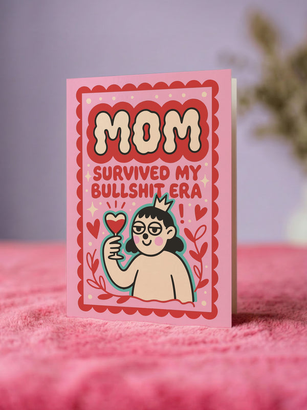 A 5x7 inch flat-printed pink greeting card featuring a cherry red scalloped border. At the top, bold wavy cream letters outlined in red read MOM. Below, red text says SURVIVED MY BULLSHIT ERA surrounded by small stars and hearts. The center illustration shows a smirking figure with black hair wearing small crown and holding a heart-shaped glass of \wine, outlined in light teal. The inside features black serif text: Bullshit era survivor award: you. Grateful you didn't quit on me. Love you, Mom.
