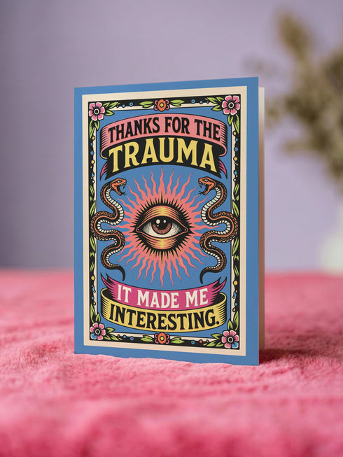 A 5x7 folded Mother's Day card with a vibrant cornflower blue background and traditional tattoo-inspired folk art. The front design is a centralized medallion featuring a realistic human eye at the center, surrounded by an orange and red radial sunburst. Two symmetrical tan and brown snakes frame the eye. Pink and yellow banners contain the black text "THANKS FOR THE TRAUMA IT MADE ME INTERESTING." Pink roses with green leaves are in corner.