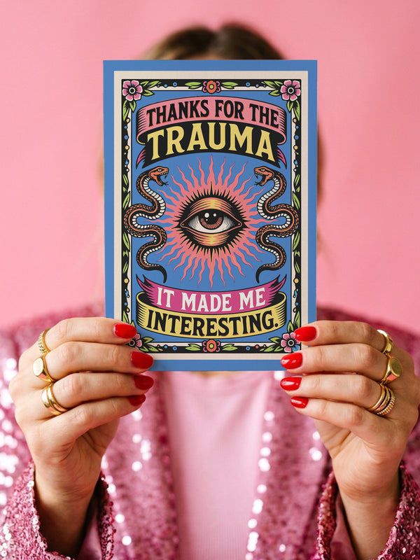 A 5x7 folded Mother's Day card with a vibrant cornflower blue background and traditional tattoo-inspired folk art. The front design is a centralized medallion featuring a realistic human eye at the center, surrounded by an orange and red radial sunburst. Two symmetrical tan and brown snakes frame the eye. Pink and yellow banners contain the black text "THANKS FOR THE TRAUMA IT MADE ME INTERESTING." Pink roses with green leaves are in corner.
