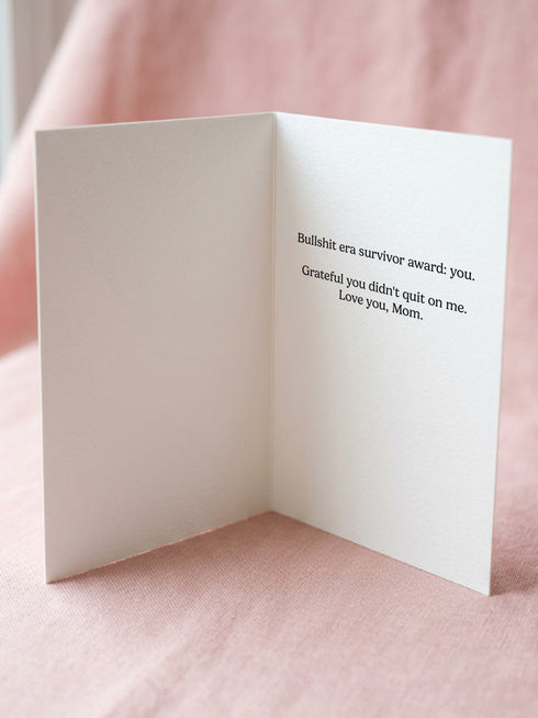 A 5x7 inch flat-printed pink greeting card featuring a cherry red scalloped border. At the top, bold wavy cream letters outlined in red read MOM. Below, red text says SURVIVED MY BULLSHIT ERA surrounded by small stars and hearts. The center illustration shows a smirking figure with black hair wearing small crown and holding a heart-shaped glass of \wine, outlined in light teal. The inside features black serif text: Bullshit era survivor award: you. Grateful you didn't quit on me. Love you, Mom.