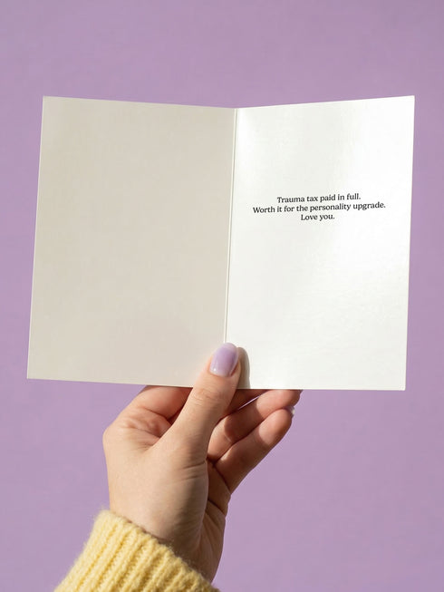 A 5x7 folded Mother's Day card with a vibrant cornflower blue background and traditional tattoo-inspired folk art. The front design is a centralized medallion featuring a realistic human eye at the center, surrounded by an orange and red radial sunburst. Two symmetrical tan and brown snakes frame the eye. Pink and yellow banners contain the black text "THANKS FOR THE TRAUMA IT MADE ME INTERESTING." Pink roses with green leaves are in corner.