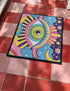 Rectangular entryway doormat with a deep purple background printed in a 2D faux embroidery illusion. The symmetrical mandala centers on a large green, pink, and white evil eye with golden yellow sunburst lines radiating outward. The corners hold textured cyan clouds, yellow crescent moons, and stars, with a scalloped floral motif along the lower edge. Finished with black trim and a non-slip rubber backing. Psychedelic celestial maximalist home decor and protective evil eye welcome mat for bold, eclectic ent