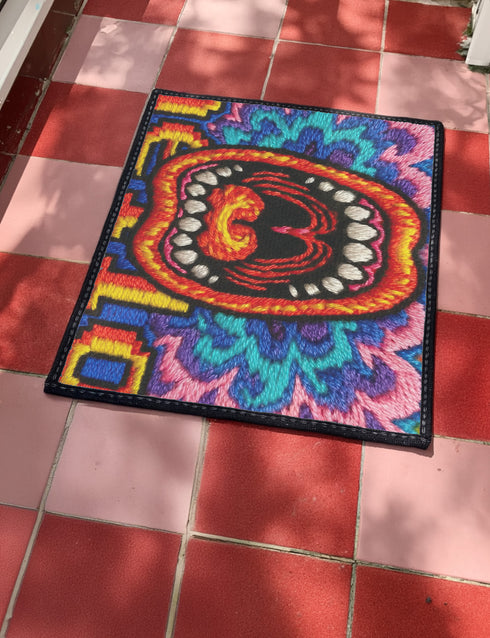 A rectangular indoor and outdoor doormat featuring a vibrant, psychedelic design. The central graphic is a large, open screaming red and orange mouth with white teeth, surrounded by highly saturated wavy, trippy patterns in blue, purple, magenta, and teal. Yellow stylized text reading HELLO is integrated into the pattern. The mat has a smooth 2D flat print surface that mimics the look of textured chainstitch embroidery, finished with a solid black fabric trim around all four outer edges.