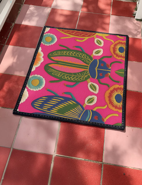 A rectangular doormat featuring a vibrant hot pink background with a high-fidelity tapestry-style print of three large, detailed beetles. The central beetle is emerald green and gold with navy accents, flanked by two side-profile beetles in ochre and deep blue. Stylized teal and yellow sunburst flowers and small sage green leaves are scattered throughout the design. The mat has a sleek black-trimmed edge and a visible flat, durable polyester texture mimicking intricate needlework.