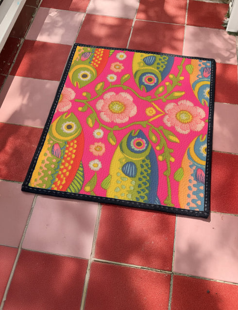 A rectangular doormat featuring a large hot pink smiley face on a deep teal background. The smiley face has green cannabis leaves for eyes and a yellow stitched-style smile. The central face is surrounded by stylized green foliage arches and concentric circles in pink, yellow, and white, resembling abstract flowers. The design mimics a vintage needlepoint or tapestry texture but is a flat print. The mat has a solid black trim border.