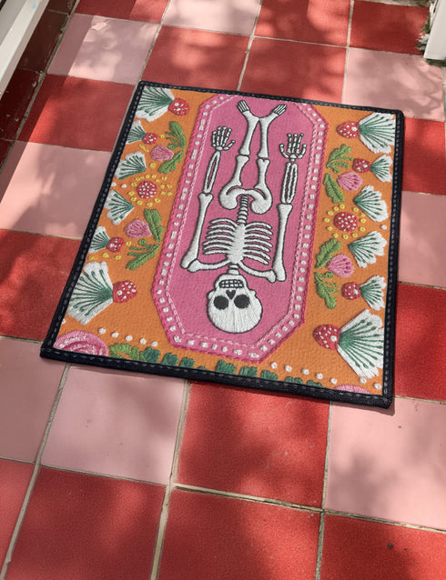 A rectangular terracotta orange doormat featuring a central illustration of a white skeleton lying inside a pink coffin shape. The design is surrounded by a symmetrical pattern of green leaves, pink flowers, and red and white spotted mushrooms. The imagery uses a trompe-l'œil effect to mimic the texture of raised needlepoint embroidery, but the surface is flat printed polyester. The mat is framed with a solid black edge.