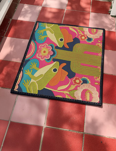 A rectangular polyester doormat with a black trim and non-slip rubber backing. The vibrant, colorful design features a symmetrical illustration on a dark navy blue background. In the center, two green frogs wearing orange cowboy hats sit facing each other. Between them is a tall, olive-green saguaro cactus. The scene is filled with large, psychedelic-style flowers in hot pink, orange, and yellow. The overall aesthetic is funky, retro 70s, maximalist, and trippy, mimicking the look of embroidery.