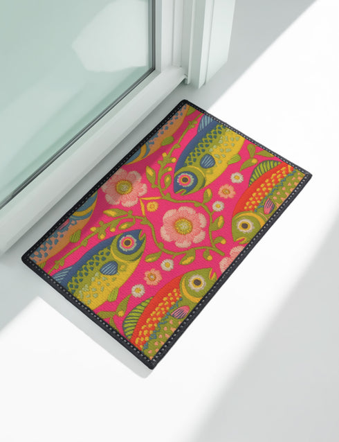 A rectangular doormat featuring a large hot pink smiley face on a deep teal background. The smiley face has green cannabis leaves for eyes and a yellow stitched-style smile. The central face is surrounded by stylized green foliage arches and concentric circles in pink, yellow, and white, resembling abstract flowers. The design mimics a vintage needlepoint or tapestry texture but is a flat print. The mat has a solid black trim border.