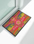 A rectangular doormat featuring a large hot pink smiley face on a deep teal background. The smiley face has green cannabis leaves for eyes and a yellow stitched-style smile. The central face is surrounded by stylized green foliage arches and concentric circles in pink, yellow, and white, resembling abstract flowers. The design mimics a vintage needlepoint or tapestry texture but is a flat print. The mat has a solid black trim border.