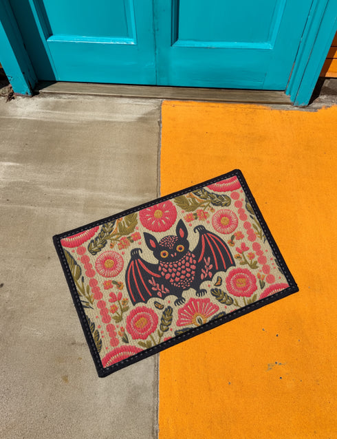 A rectangular beige doormat featuring a large, symmetrical folk art bat illustration in the center. The bat is black with coral red and mustard gold details that mimic the look of satin stitch embroidery. Surrounding the bat are stylized floral motifs in deep pink, coral, and olive green, arranged in a dense, decorative pattern. The texture visually resembles woven linen but is a smooth, flat print. The mat has a solid black trim border and is shown on a flat surface.