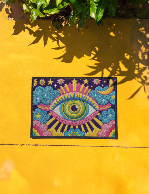 Rectangular entryway doormat with a deep purple background printed in a 2D faux embroidery illusion. The symmetrical mandala centers on a large green, pink, and white evil eye with golden yellow sunburst lines radiating outward. The corners hold textured cyan clouds, yellow crescent moons, and stars, with a scalloped floral motif along the lower edge. Finished with black trim and a non-slip rubber backing. Psychedelic celestial maximalist home decor and protective evil eye welcome mat for bold, eclectic ent