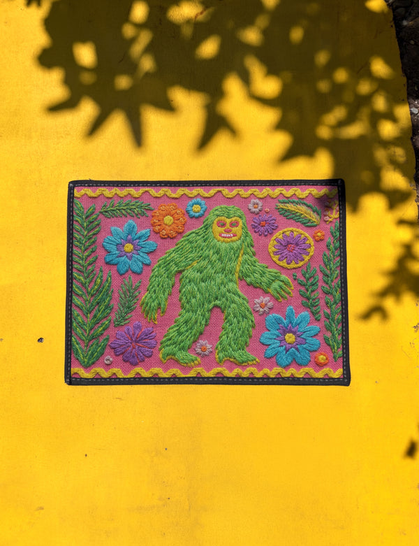 A rectangular flat-printed doormat featuring a faux-tufted, trompe-l'œil texture design. The background is a vibrant hot pink, bordered by a wavy yellow line. In the center is a bright green, smiling Bigfoot or Sasquatch figure with long shaggy hair. Surrounding the cryptid are highly detailed, colorful faux-embroidered floral motifs including blue, orange, and purple daisies, alongside green fern fronds and leaves. The mat is finished with a solid black fabric trim around the outer edge.